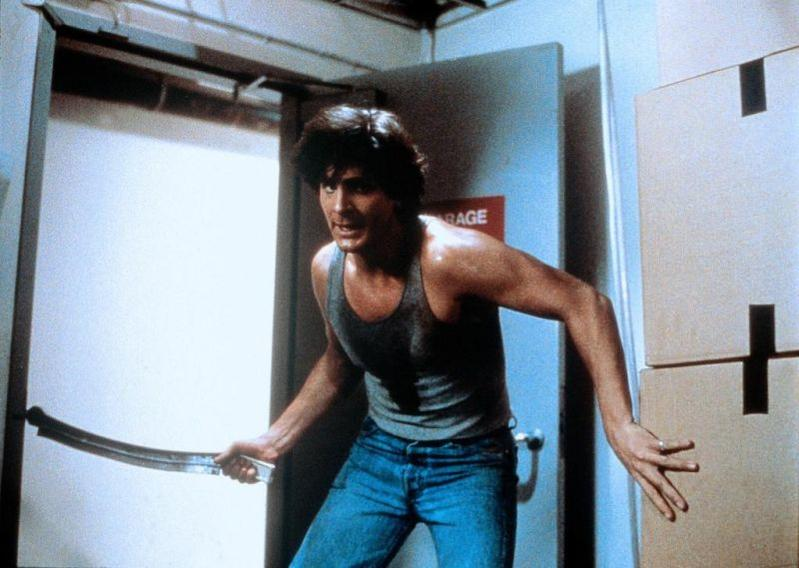 Mann in grauem Tanktop und Jeans hält gebogenes Metallstück, steht vor Lagerräumen.