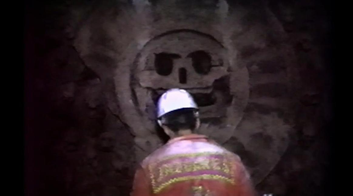 Ein Mann in einem weißen Helm blickt auf ein Totenkopf-Relief an einer Wand.