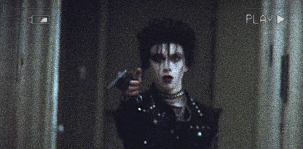 Eine Person mit Gothic-Make-up und dunkler Kleidung hält eine Pistole.