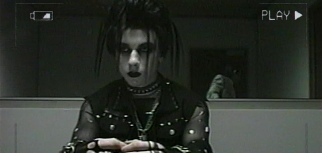Ein Goth-Jugendlicher mit dunklem Make-up sitzt und trägt eine Halskette und eine Weste.