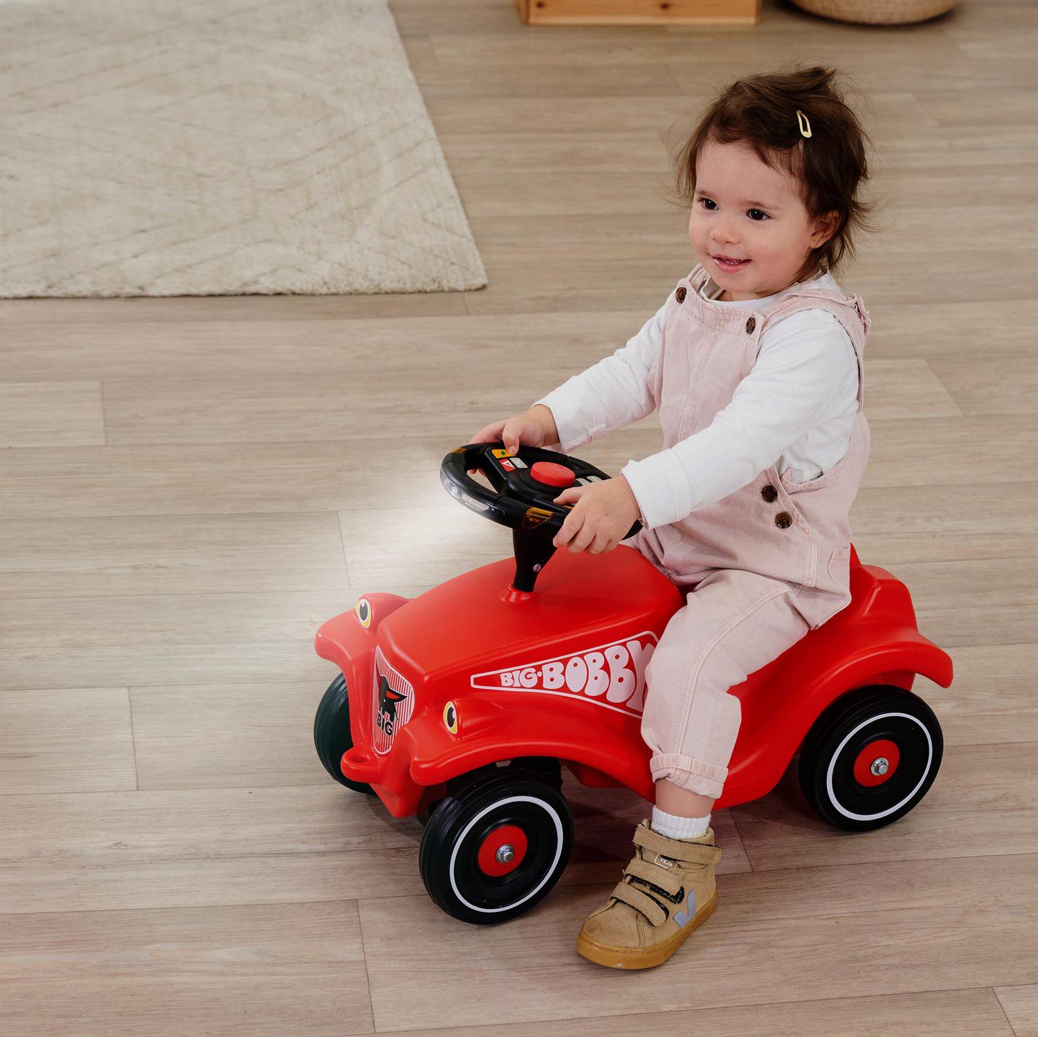 Ein Baby sitzt auf einem roten Spielzeugauto mit Lenkrad und lächelt.