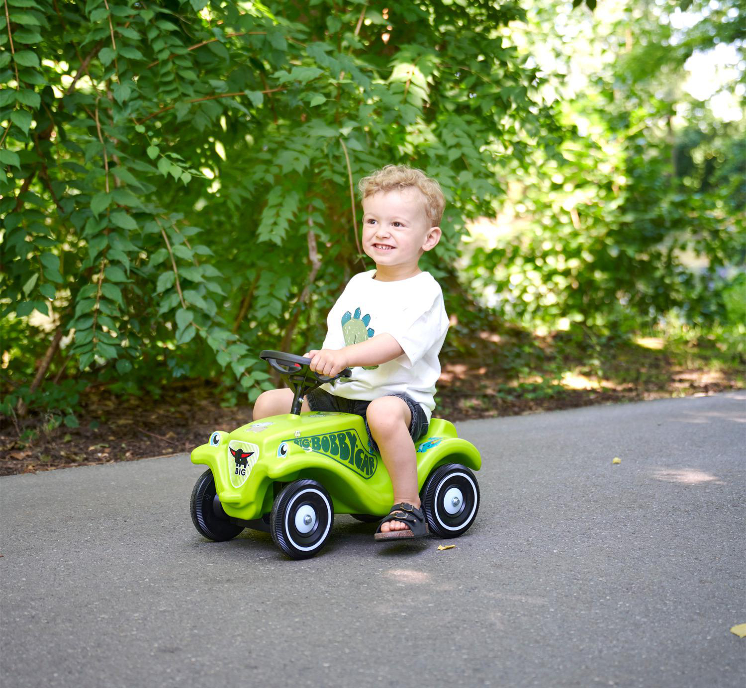 Ein kleiner Junge fährt ein grünes Bobby-Car auf einem Weg im Freien.