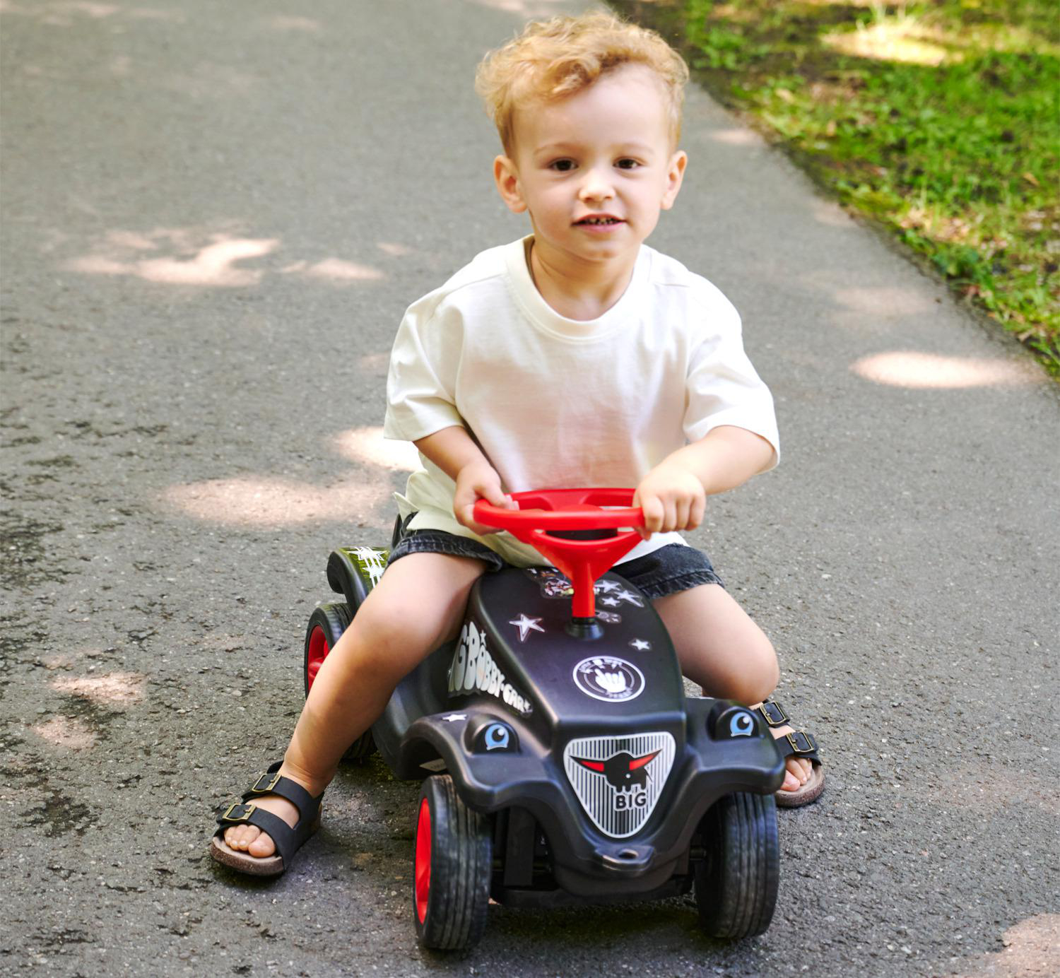 Ein kleiner Junge mit blonden Haaren sitzt auf einem schwarzen Spielzeugauto mit rotem Lenkrad im Freien.