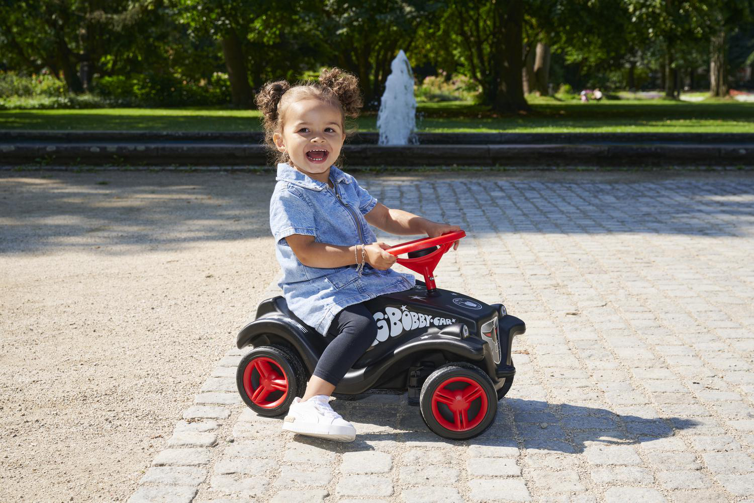 Kleines Mädchen mit Duttfrisur sitzt auf einem schwarzen Bobby-Car mit roten Rädern im Park.