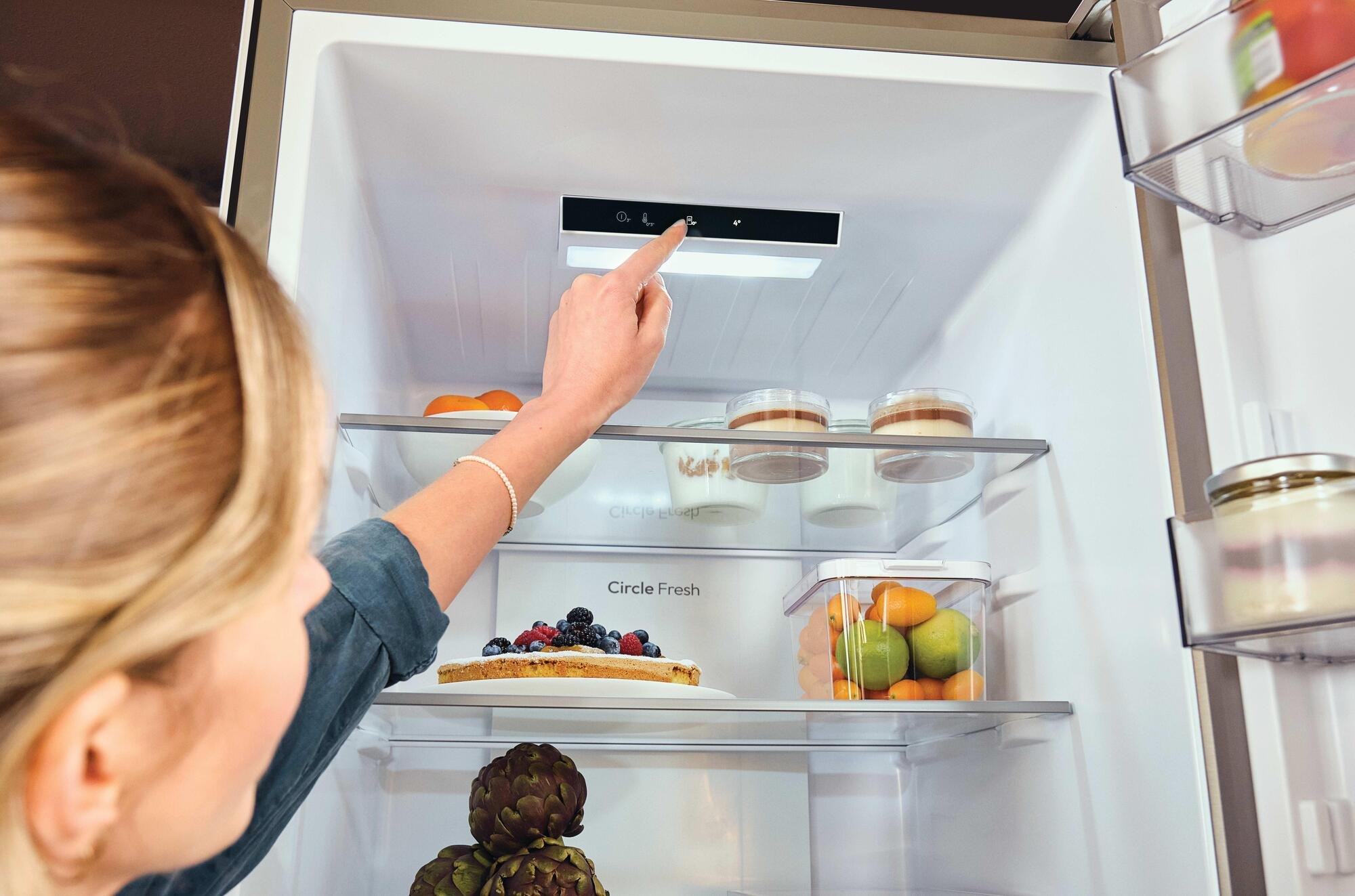 Una mano presiona un panel de control dentro de un refrigerador, con alimentos cerca.