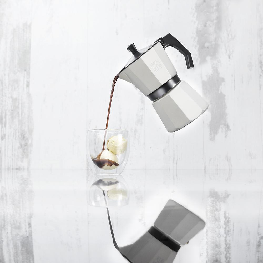 Una cafetera Moka vierte café en un vaso con helado sobre un espejo.