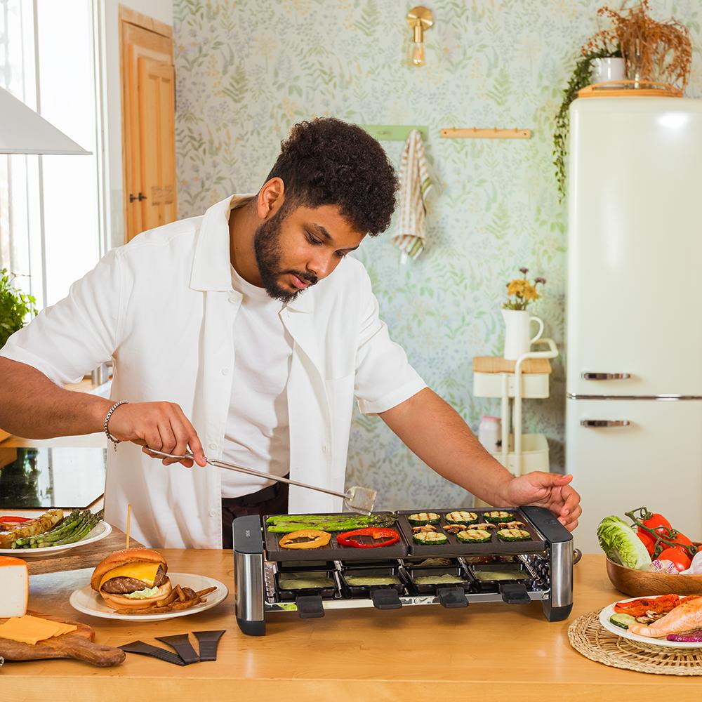 Hombre cocina verduras en parrilla raclette con hamburguesa y salmón.