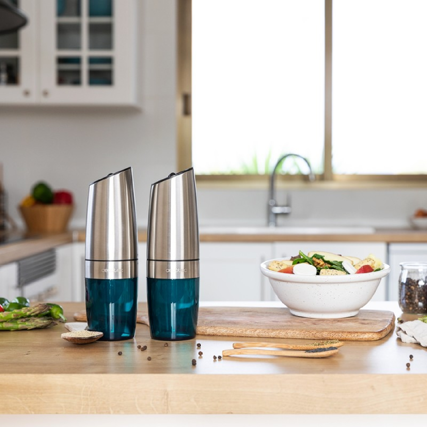 Dos molinillos de acero inoxidable con bases de cristal azul están sobre un mostrador de madera cerca de una ensalada.