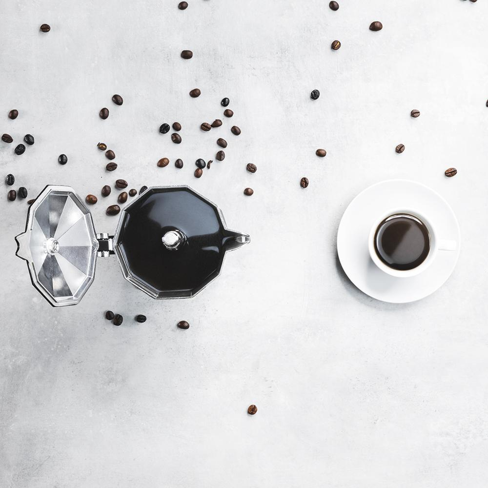 Una cafetera moka y una taza de café sobre una superficie clara con granos de café.