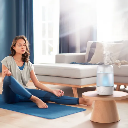 Egy nő meditál kék szőnyegen egy világos szobában, diffúzorral.
