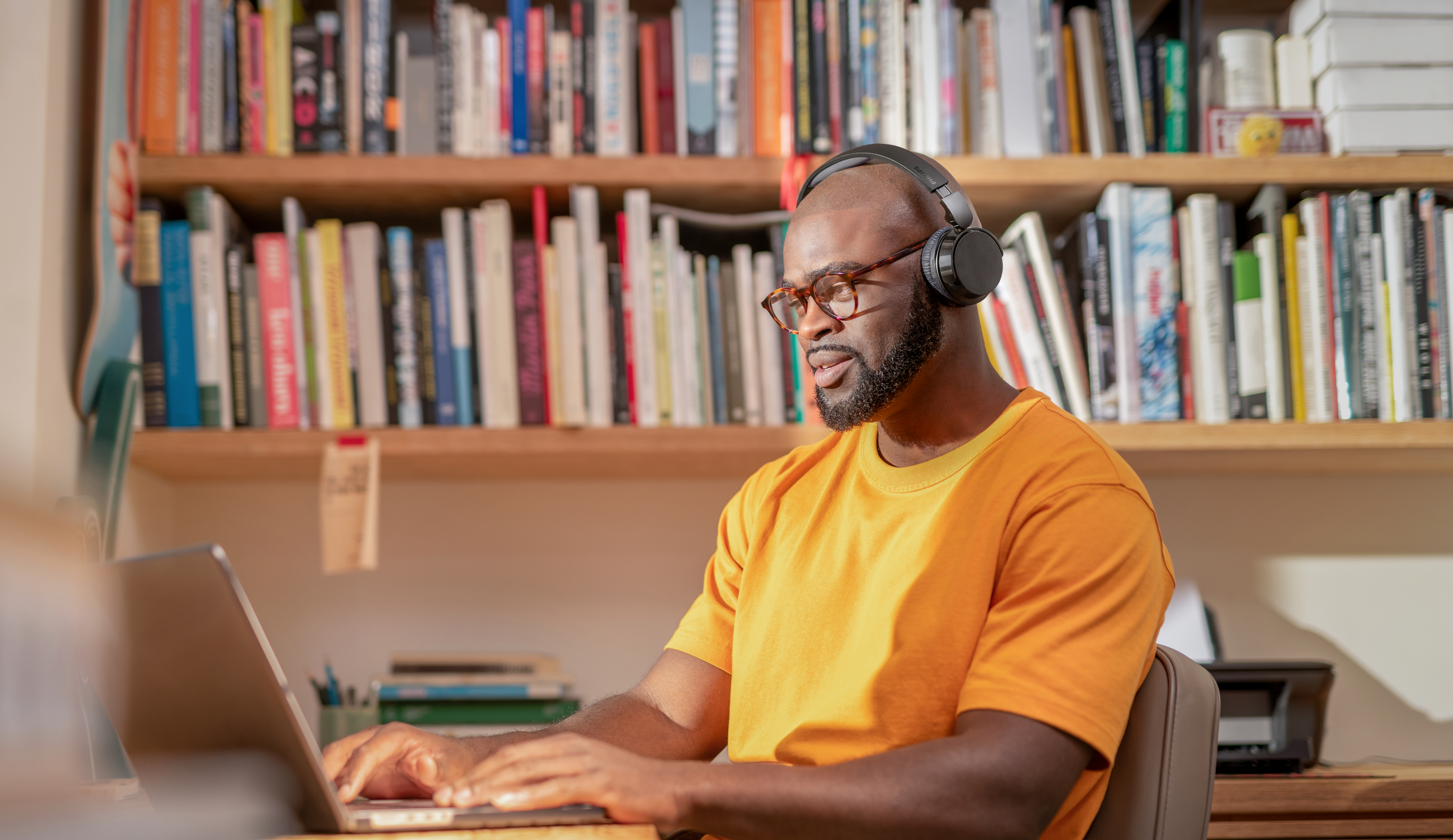 Mann mit Kopfhörern tippt auf Laptop vor Bücherregal.