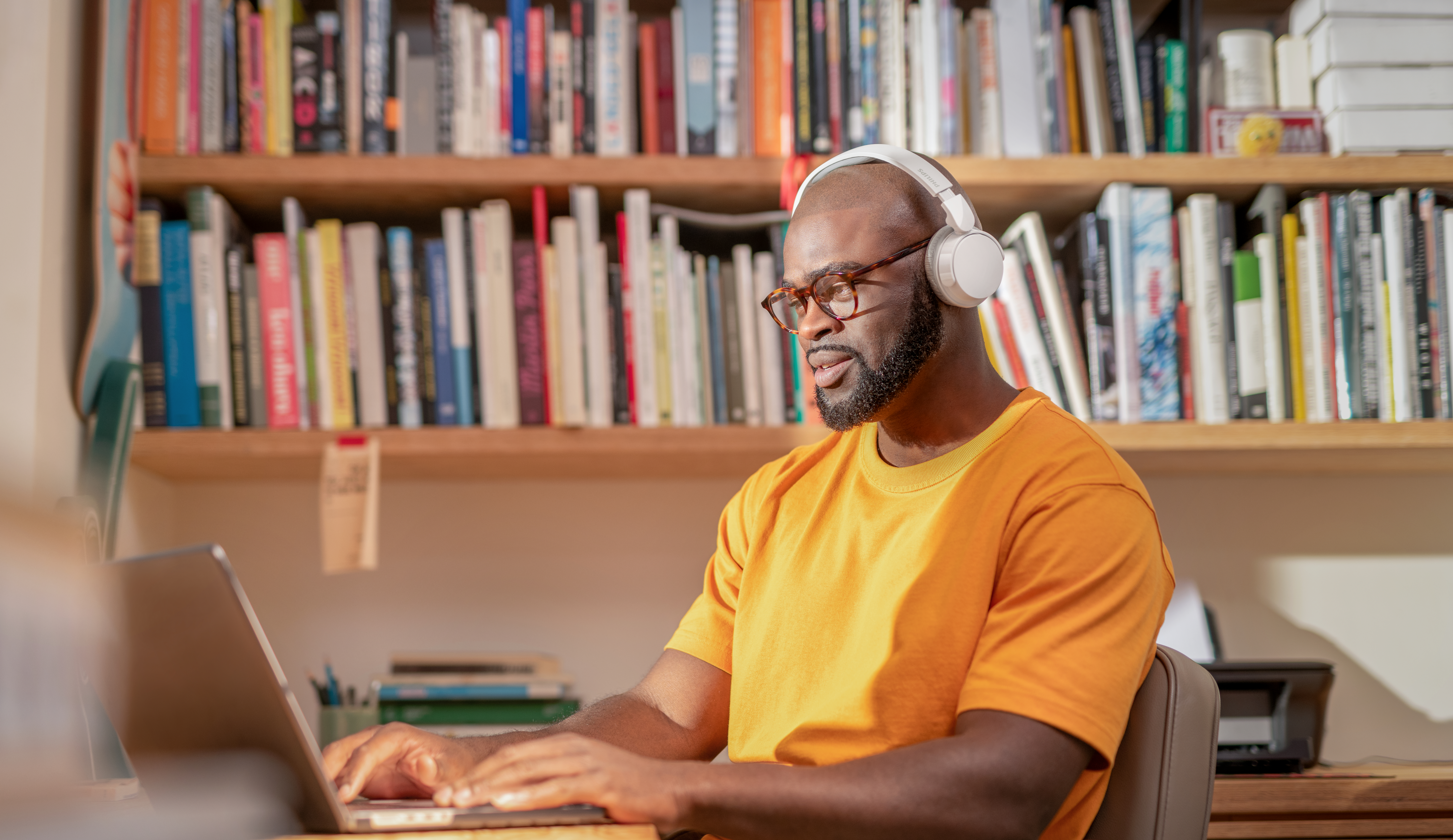 Mann mit Kopfhörern und Brille tippt auf einem Laptop vor einem Bücherregal.