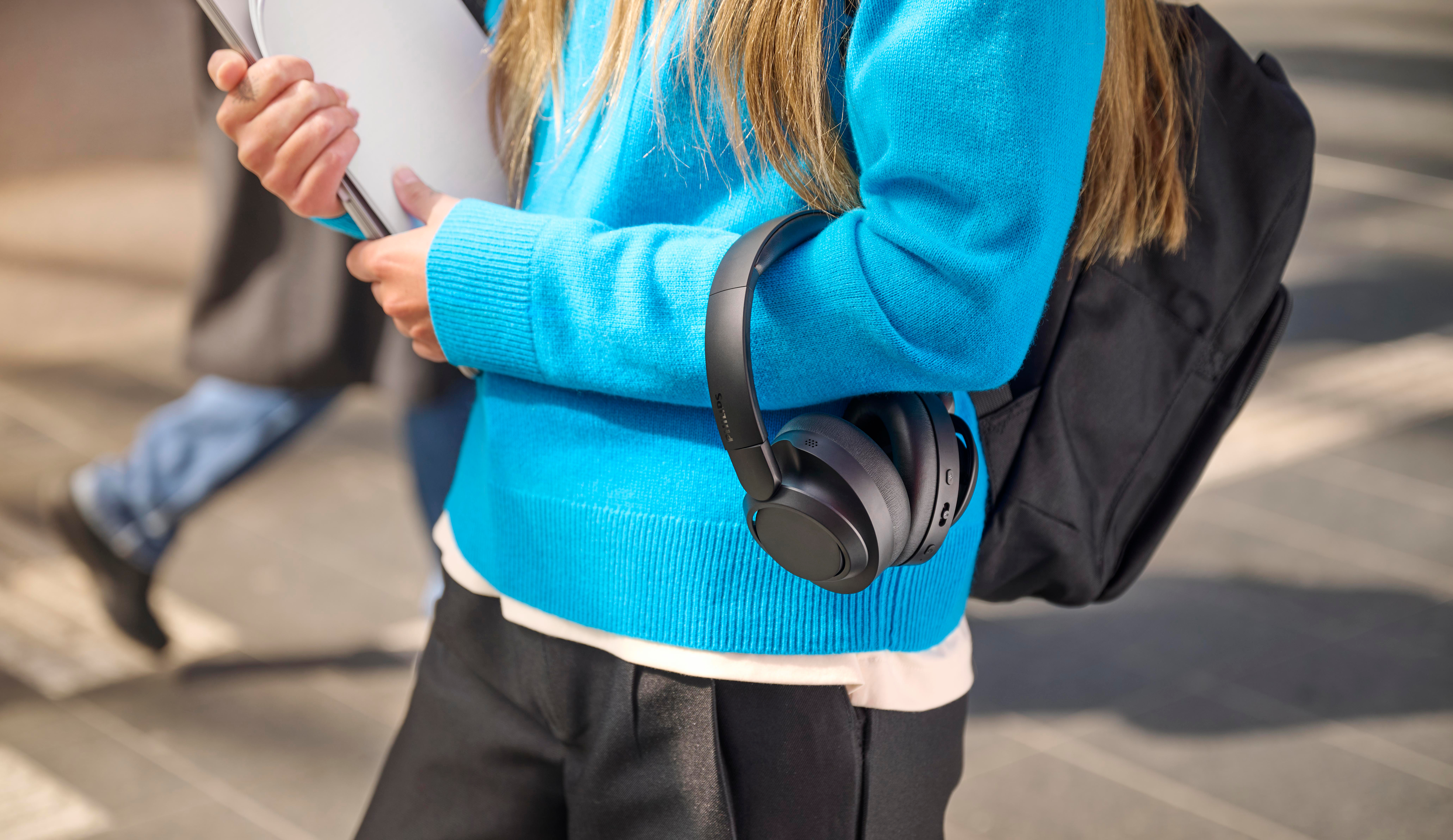 Person mit blauem Pullover, Kopfhörern, Rucksack und Laptop im Freien.