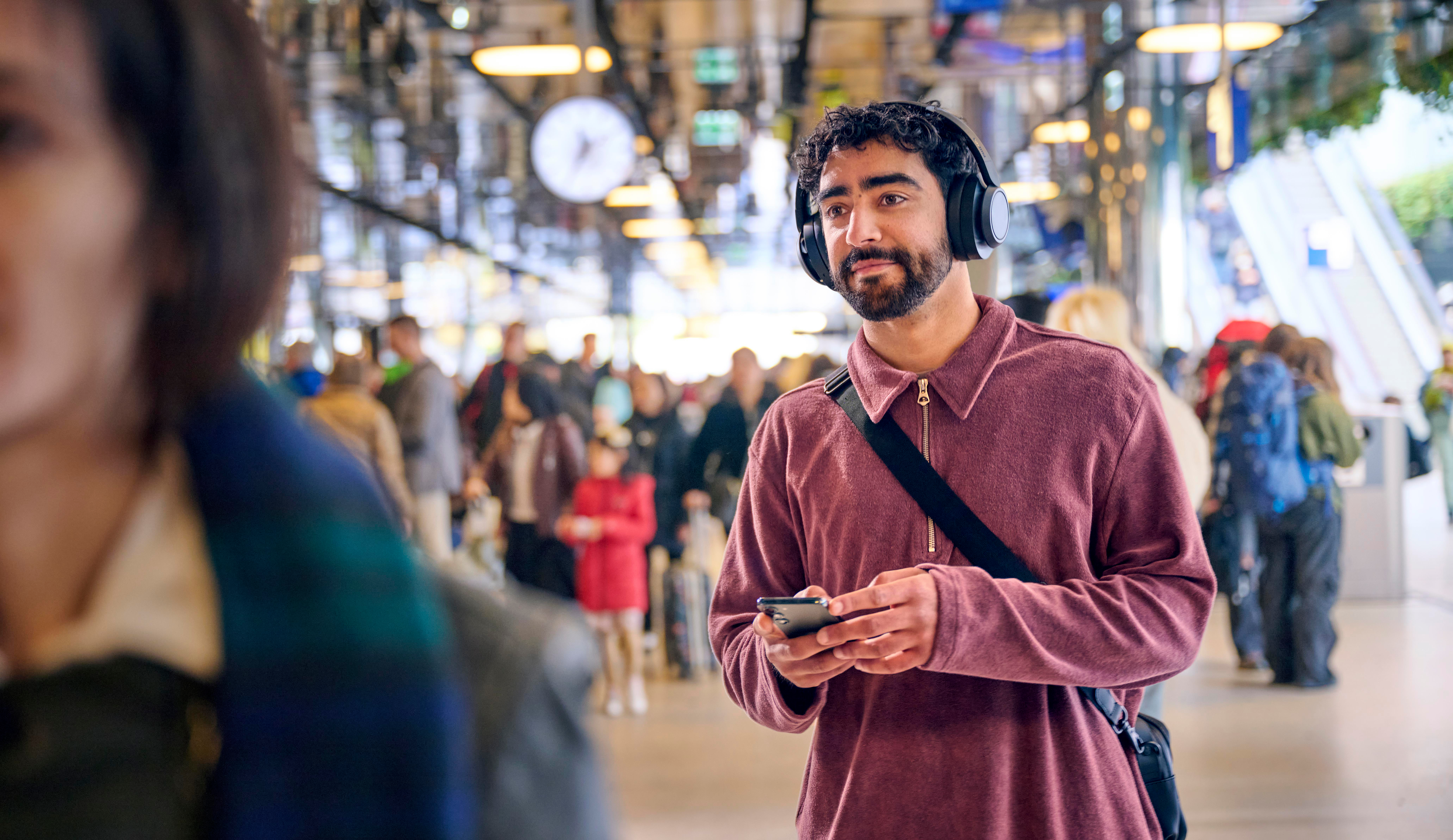 Mann mit Kopfhörern und Handy in belebtem Bahnhof, schaut nach rechts.