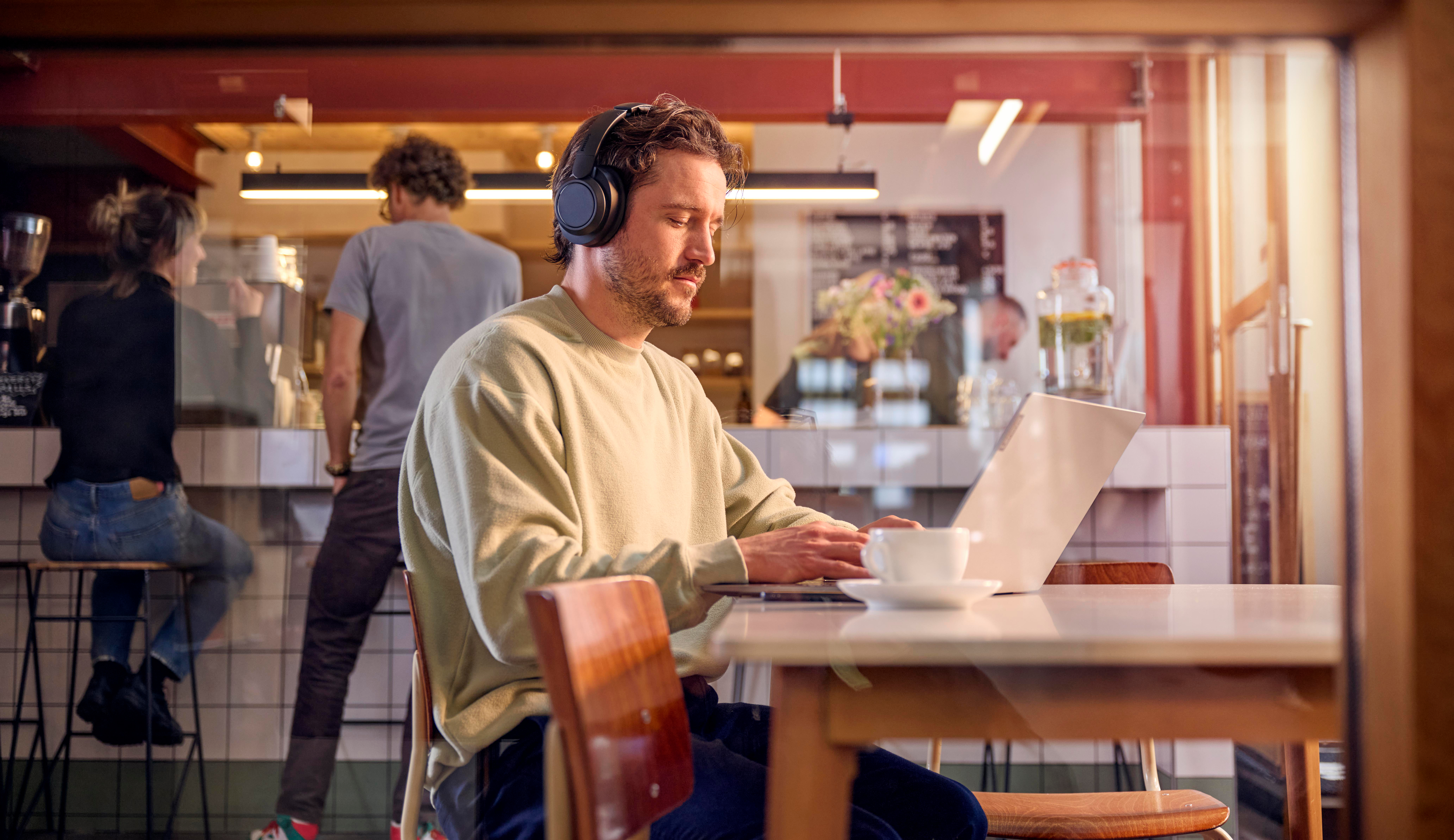 Mann mit Kopfhörern arbeitet am Laptop in einem Café. Im Hintergrund sind Gäste.