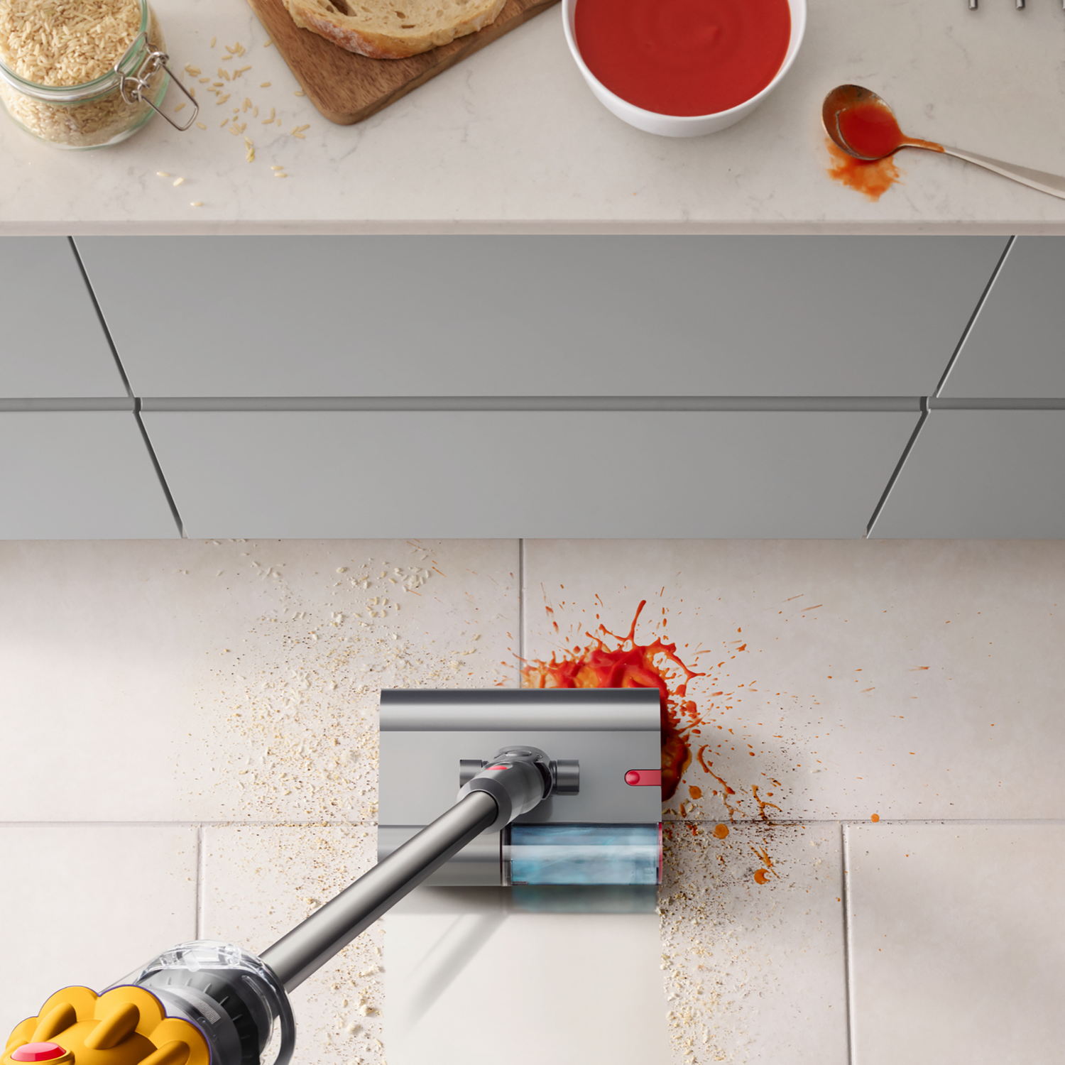 A vacuum cleaner cleans rice and tomato splashes on a light tile floor.
