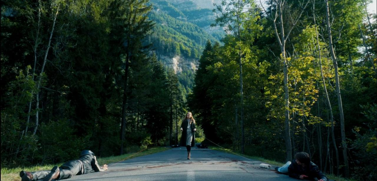 Frau geht auf Straße, zwei Personen liegen am Boden, Wald und Berge im Hintergrund.