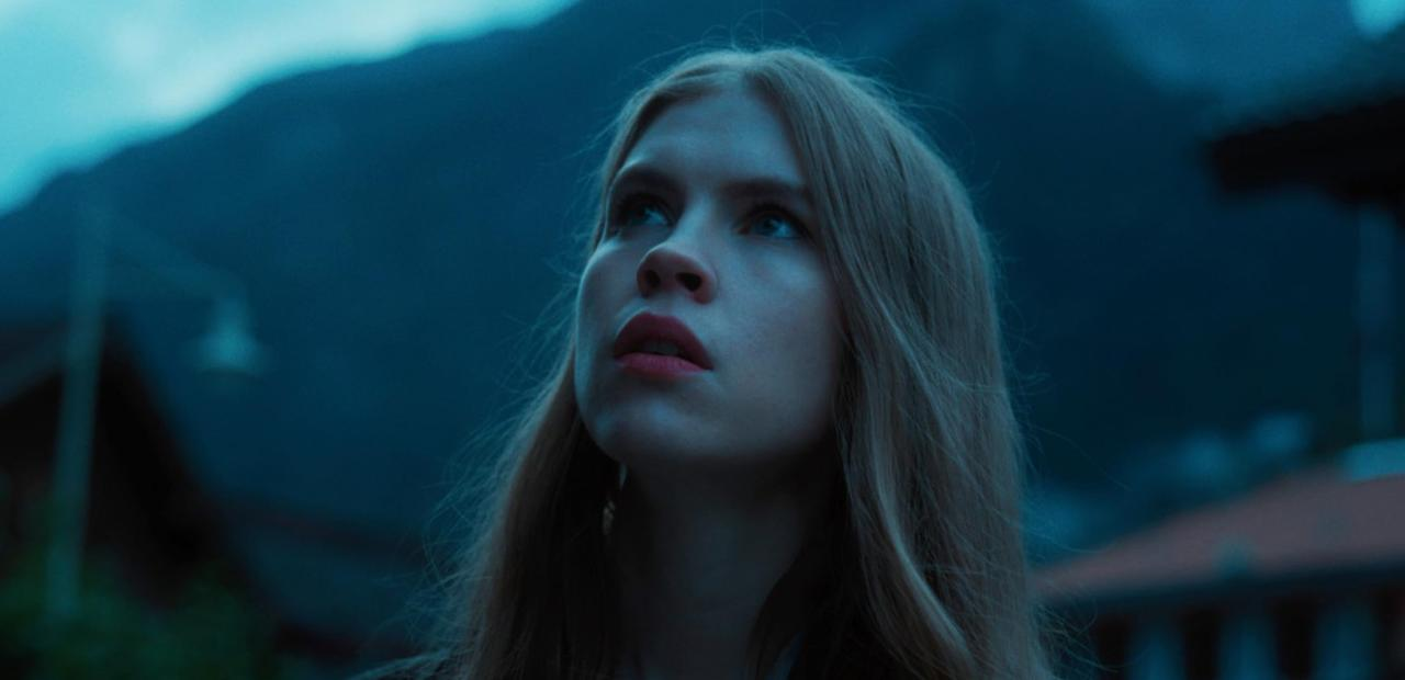 Eine junge Frau mit blonden Haaren blickt nach oben in eine dunkle, bergige Landschaft.