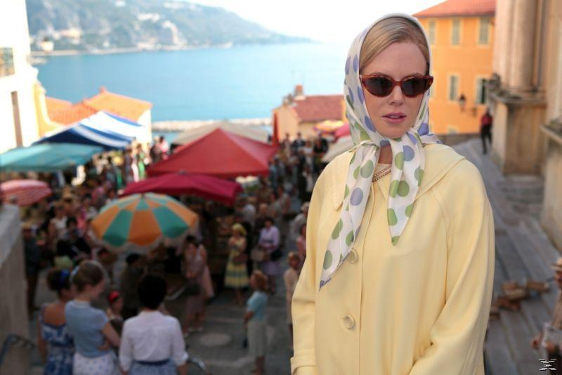 Eine Frau in gelber Jacke und Kopftuch steht vor einem Markt und dem Meer.