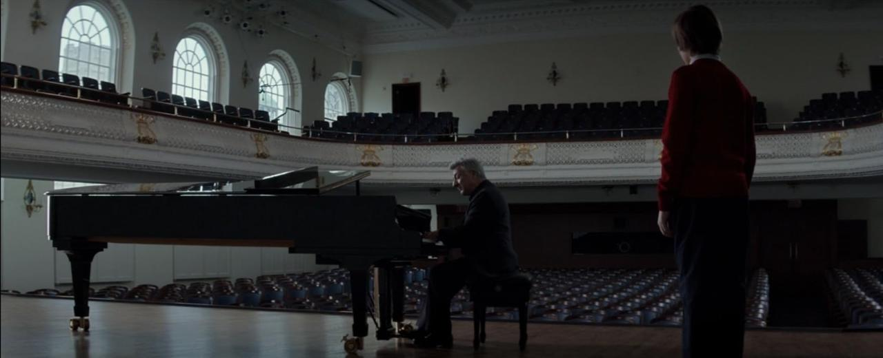 Ein Mann spielt Klavier in einem leeren Konzertsaal, ein Junge steht.