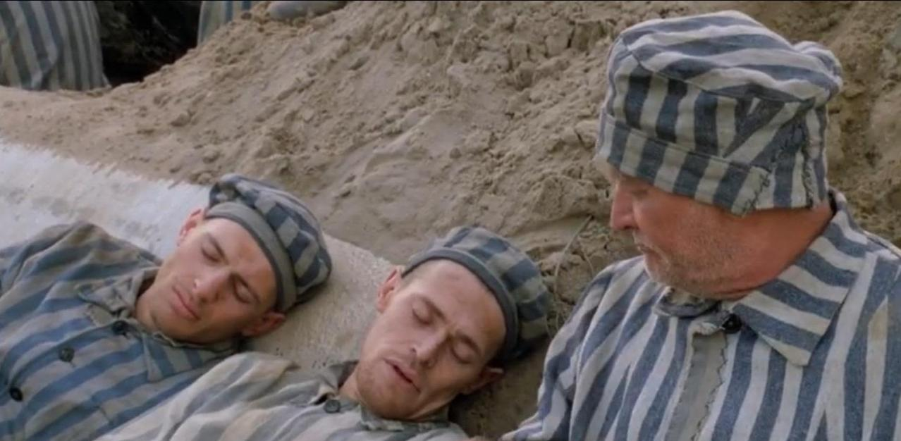 Drei Männer in gestreiften Gefängnisuniformen liegen oder sitzen im Freien.