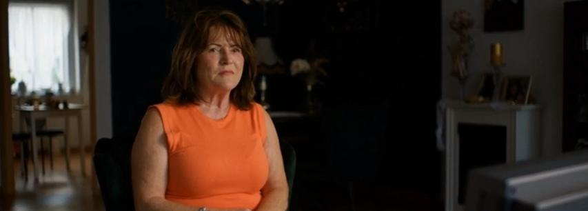 Eine Frau mittleren Alters in einem orangefarbenen Oberteil sitzt in einem dunklen Raum.