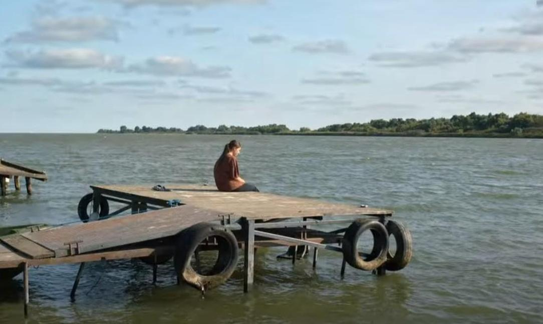 Eine Person sitzt auf einem Holzsteg am Wasser. Reifen sind am Steg befestigt.