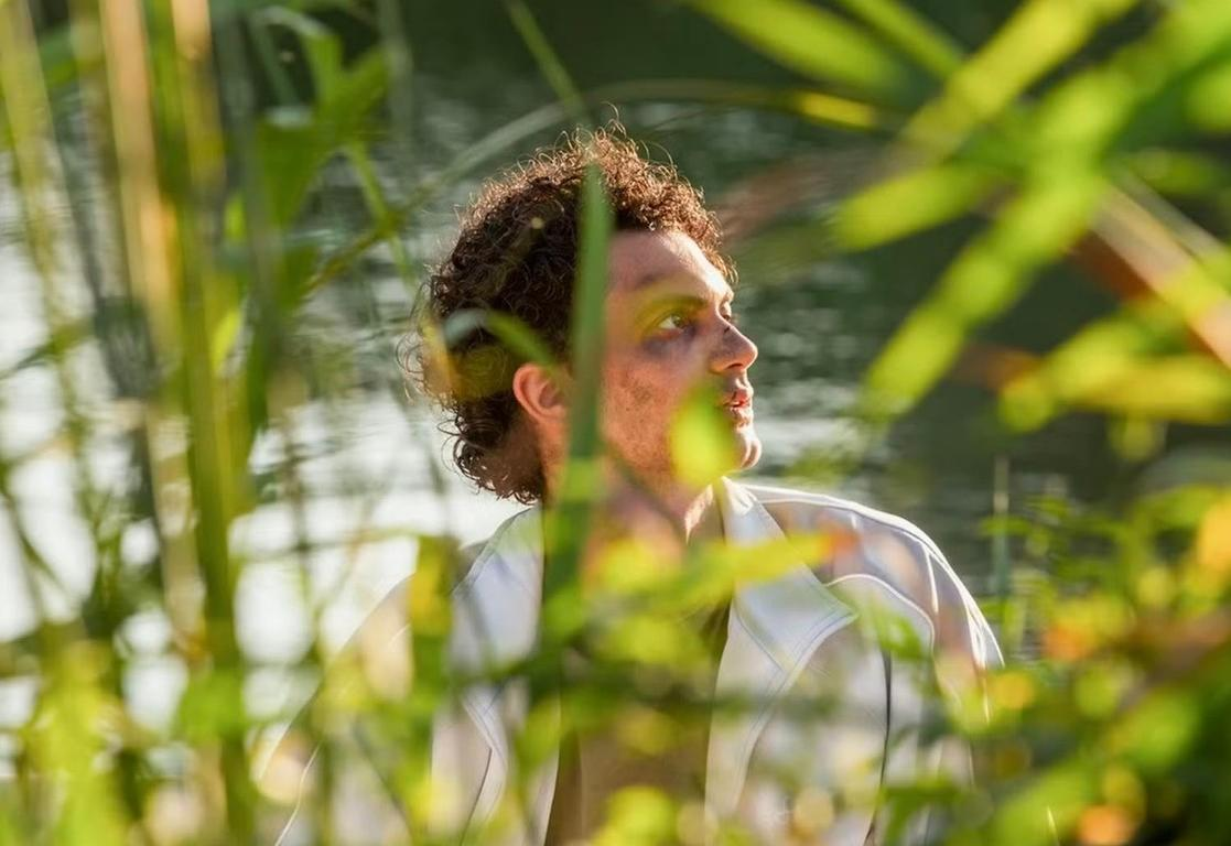 Mann mit Locken blickt nach rechts, durch grünes Laub gefiltert.