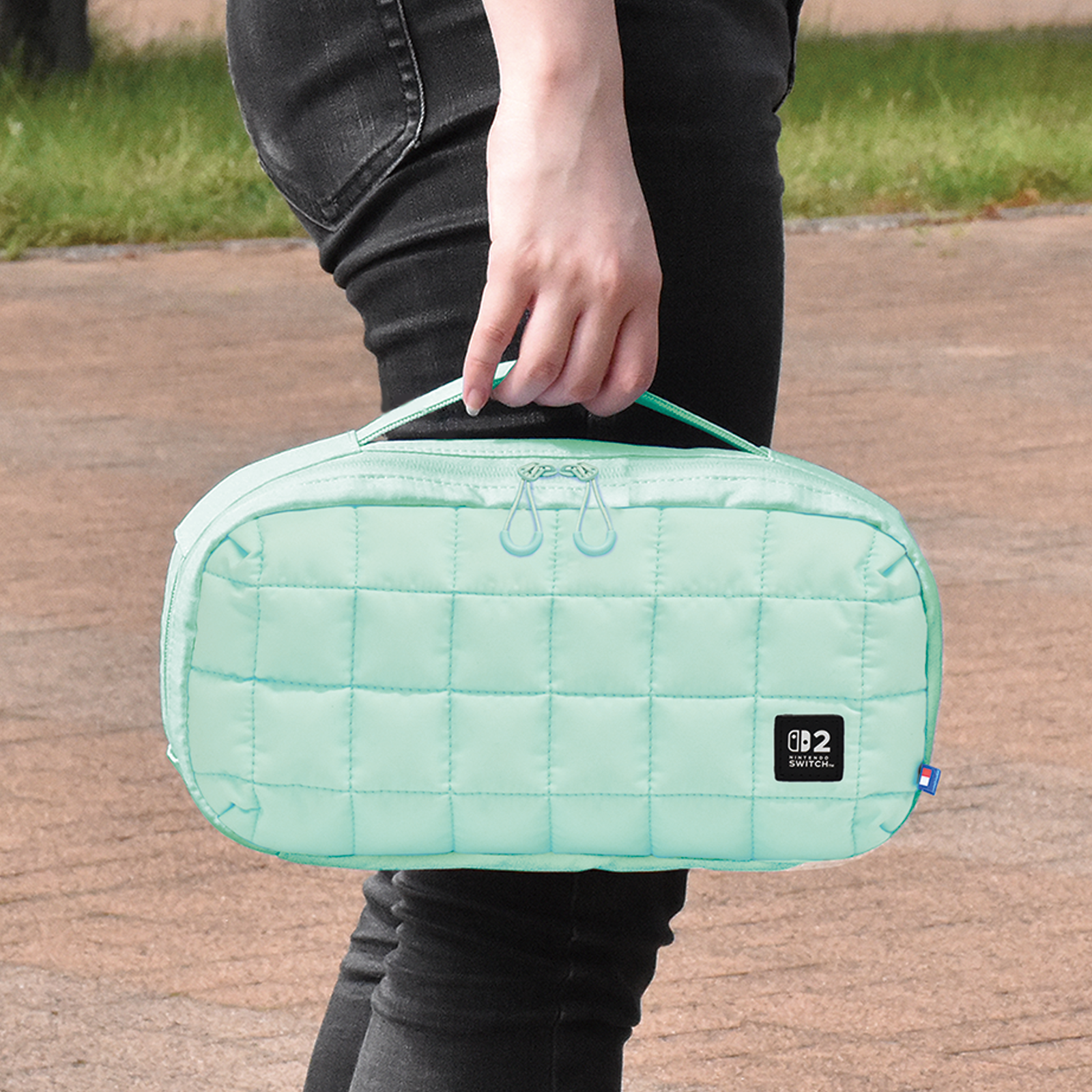 Eine Person hält eine gesteppte mintgrüne Tasche mit zwei Reißverschlüssen und einem Nintendo Switch-Logo.