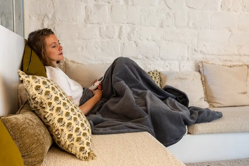 Una donna è sdraiata su un divano sotto una coperta grigia, con in mano un telecomando.