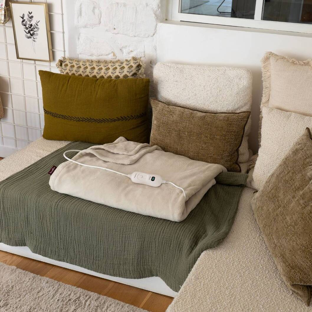 Una coperta riscaldata beige giace su una coperta verde con cuscini sullo sfondo.