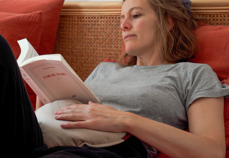 Une femme est allongée sur un canapé, lisant un livre. Elle a une bouillotte sur le ventre.