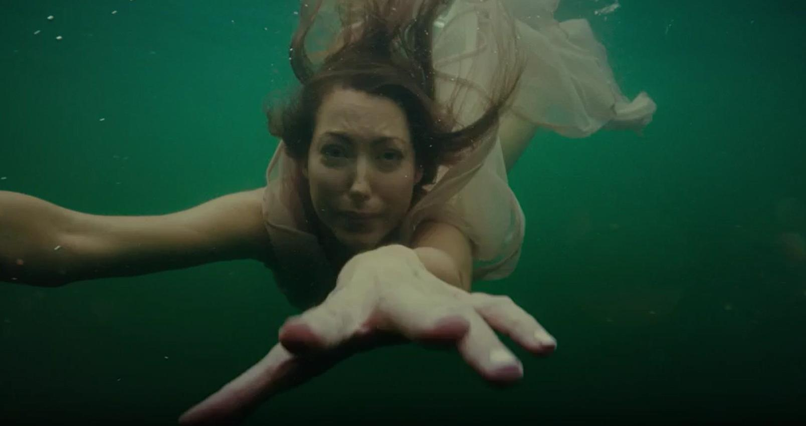 Eine Frau in einem hellen Kleid schwimmt unter Wasser, ihr Gesicht ist besorgt.