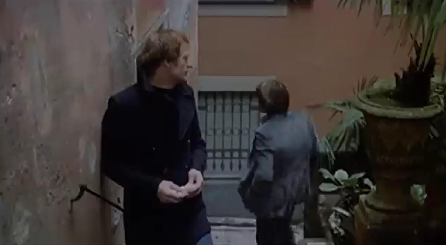 Zwei Männer stehen vor einer Wand. Einer trägt einen dunklen Mantel.