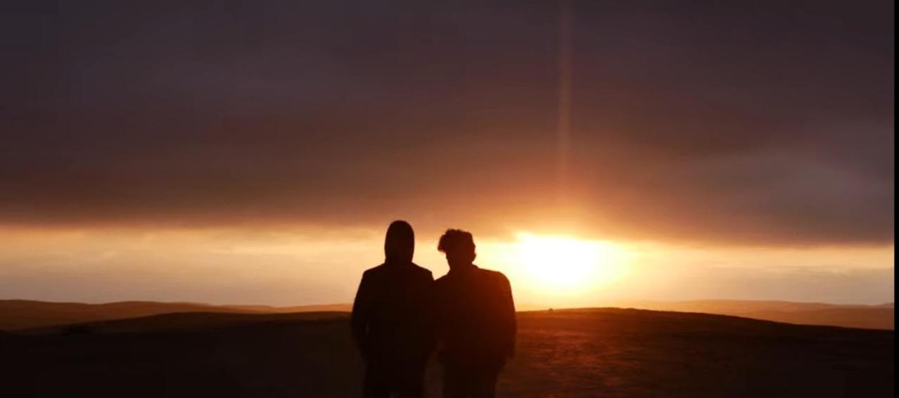 Zwei Silhouetten stehen auf Hügeln bei Sonnenuntergang, orangefarbener Himmel.
