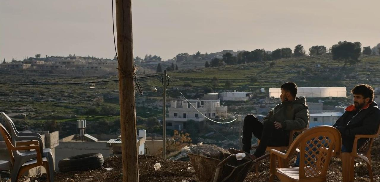 Zwei Männer sitzen auf Stühlen mit Blick auf eine hügelige Landschaft mit Gebäuden.
