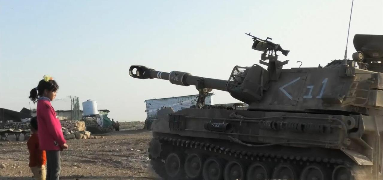 Ein Mädchen in einem rosafarbenen Pullover steht vor einem Panzer in einer ländlichen Gegend.