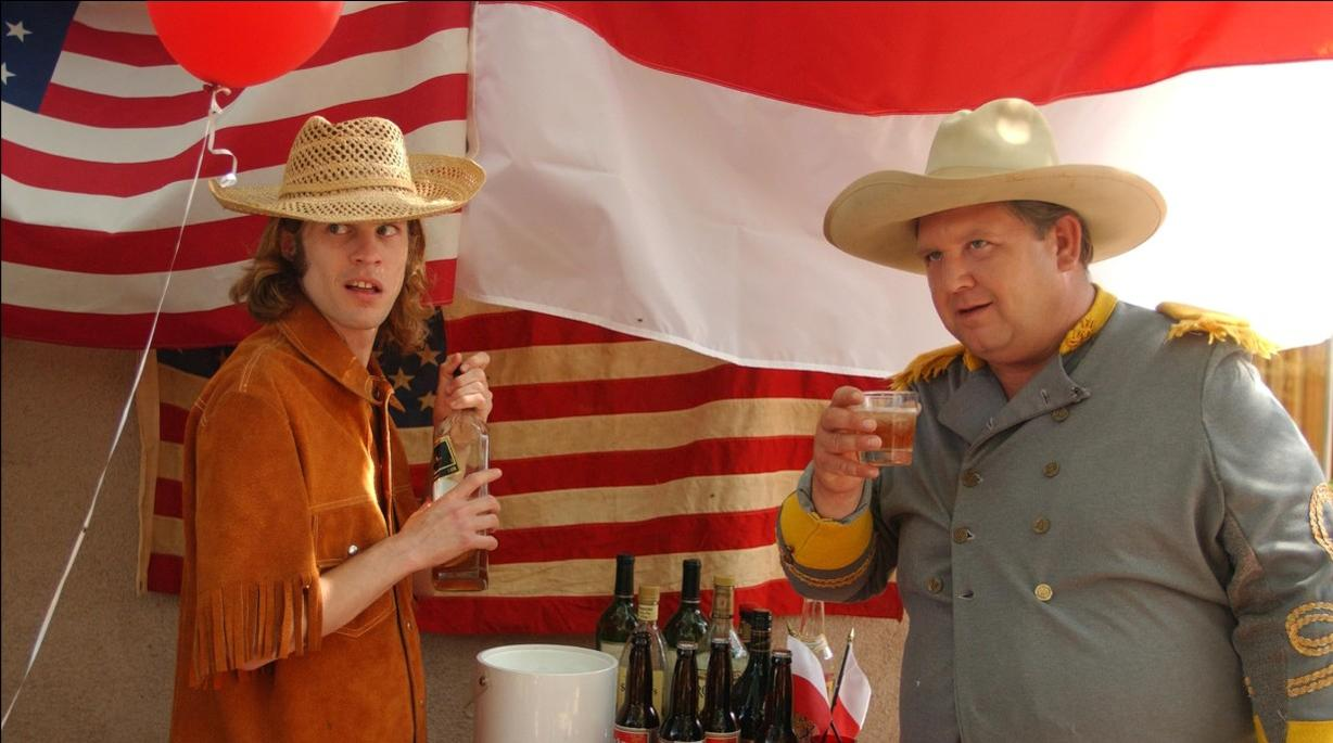 Zwei Männer in Hüten stehen vor einer US-Flagge. Einer trägt ein braunes Hemd, der andere eine graue Uniform.