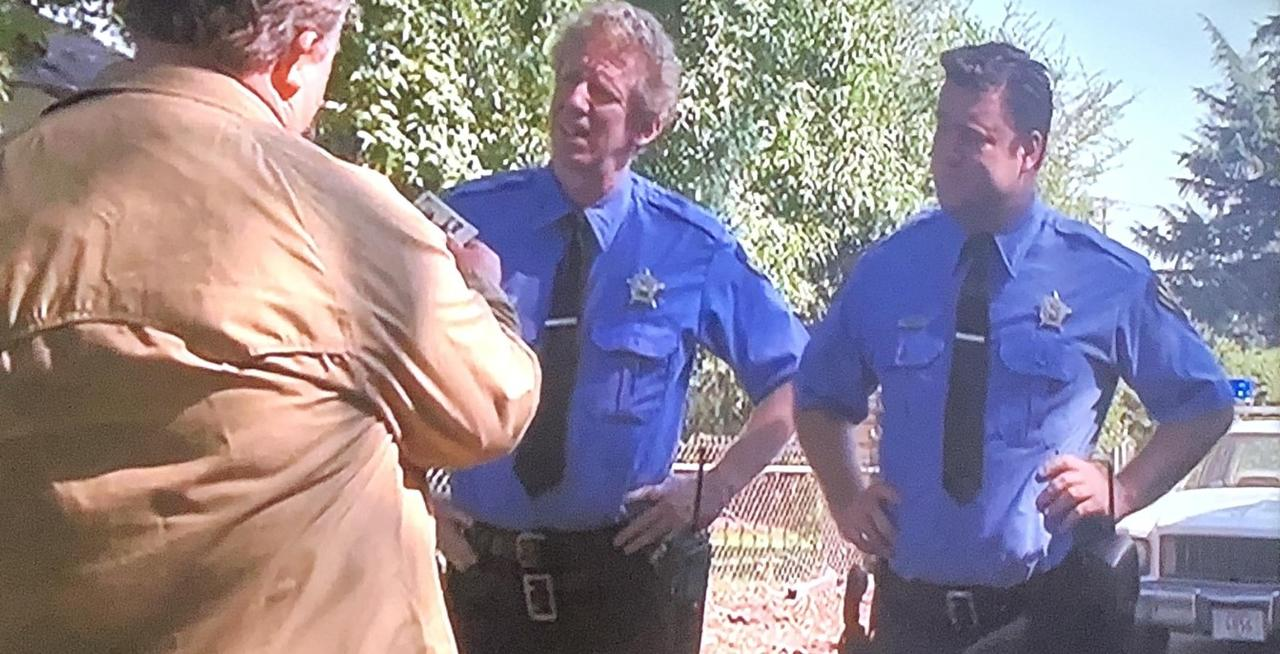 Drei Männer sprechen im Freien. Zwei Polizisten in blauen Uniformen und ein Mann in einem beigen Mantel.