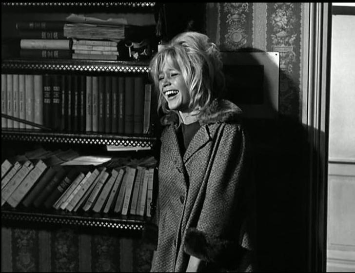 Eine lachende Frau mit blonden Haaren steht vor einem Bücherregal.