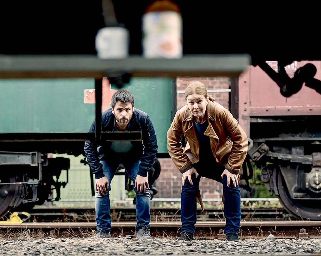 Mann und Frau kauern auf Bahngleisen vor einem Zug. Mann trägt dunkle Jacke, Frau braune.