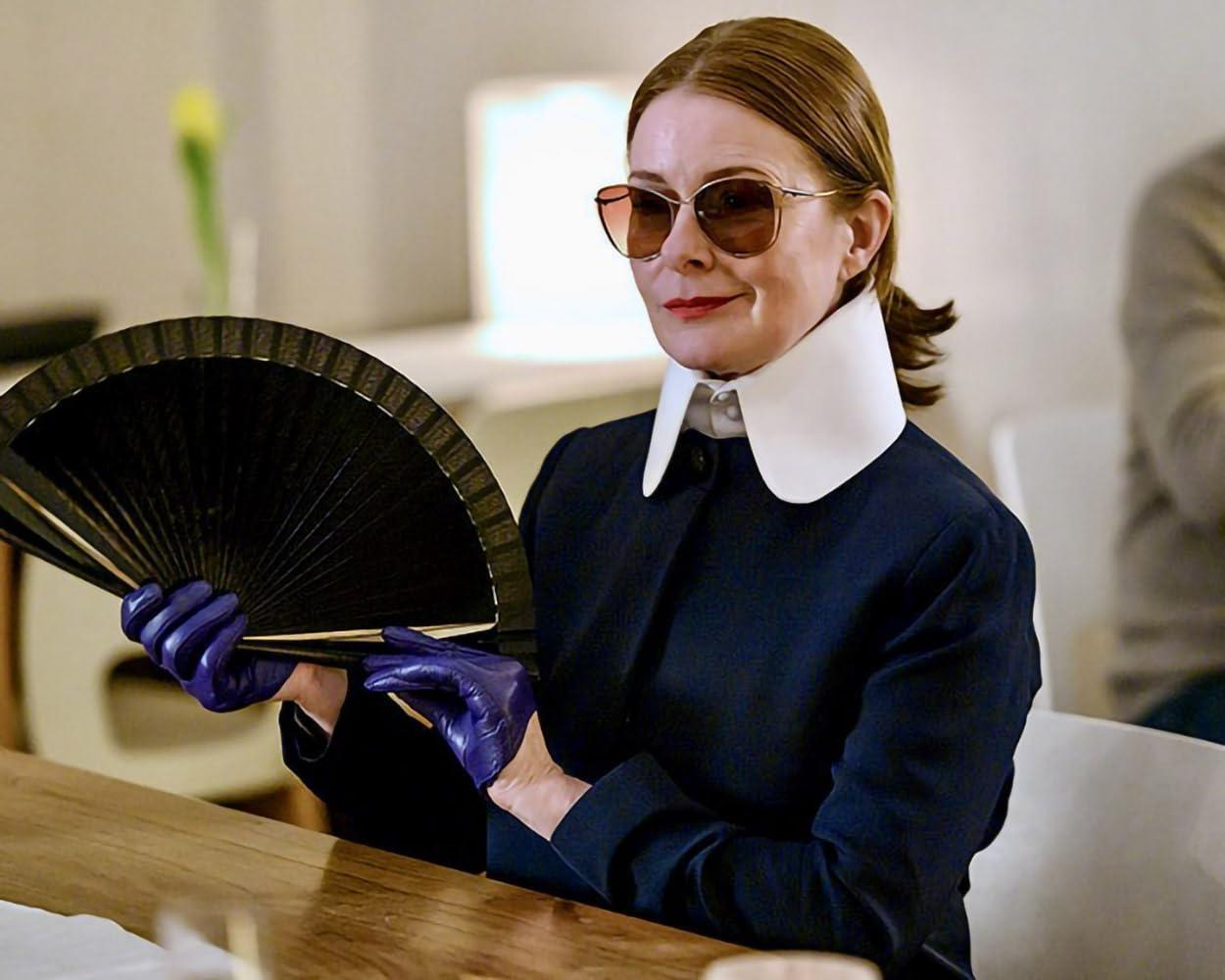 Eine Frau mit Sonnenbrille hält einen schwarzen Fächer und trägt blaue Handschuhe.