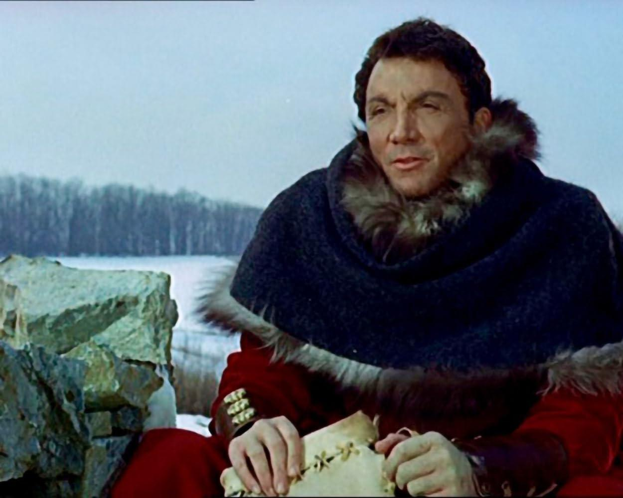 Ein Mann mit Pelzmantel und Kapuze sitzt vor einer verschneiten Landschaft.
