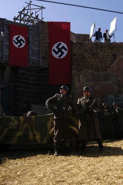 Zwei Soldaten in Uniformen stehen vor einem Fahrzeug. Rote Fahnen mit Hakenkreuzen hängen.