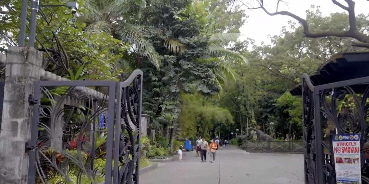 Eingang zu einem Park mit Bäumen, Toren und einigen Besuchern.
