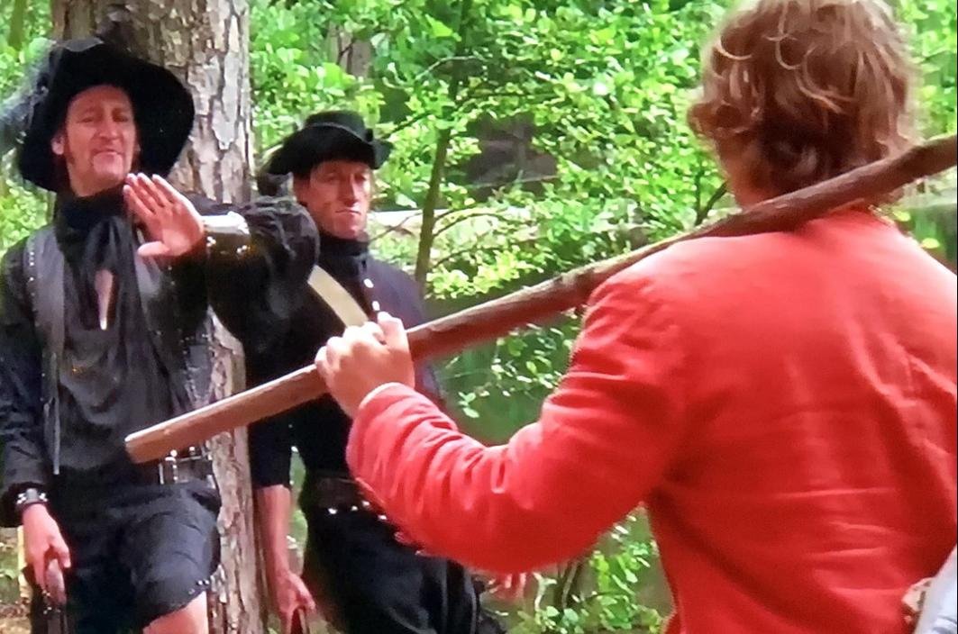 Drei Männer in historischer Kleidung im Wald. Einer trägt Rot, zwei tragen Schwarz.