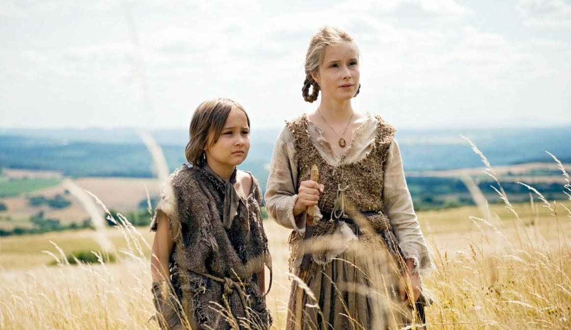 Zwei Kinder in rustikaler Kleidung stehen auf einem Feld mit hohem, trockenem Gras. Eine ferne, dunstige Landschaft.