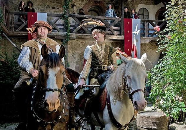 Zwei Männer in historischer Kleidung reiten auf Pferden vor einer Burg.