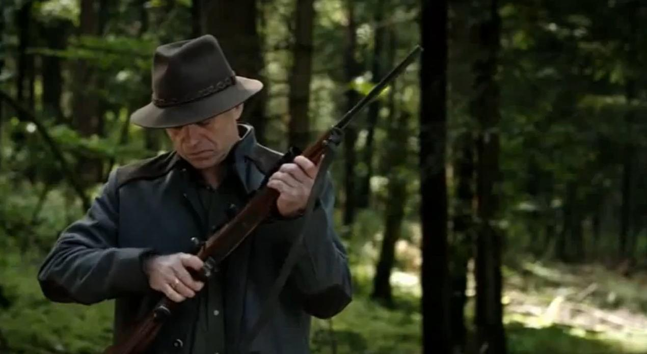Ein Mann mit Hut und Jagdjacke hält ein Gewehr im Wald.