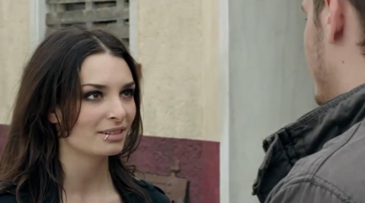 Eine Frau mit dunklem Haar und Piercing blickt nach rechts, ein Mann ist im Hintergrund.